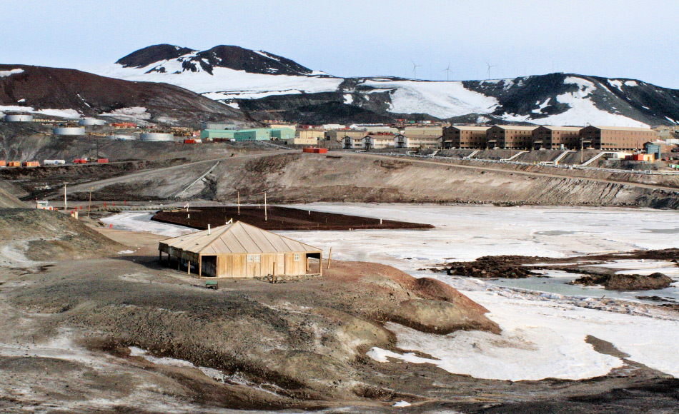 Die Discovery-HÃ¼tte auf Ross Island und im Hintergrund die US-amerikanische Basis McMurdo. Von hier aus startete Scott sein Wissenschafts- und Entdeckungsprogramm bis tief in den Kontinent hinein. Bild: Taylor & Francis